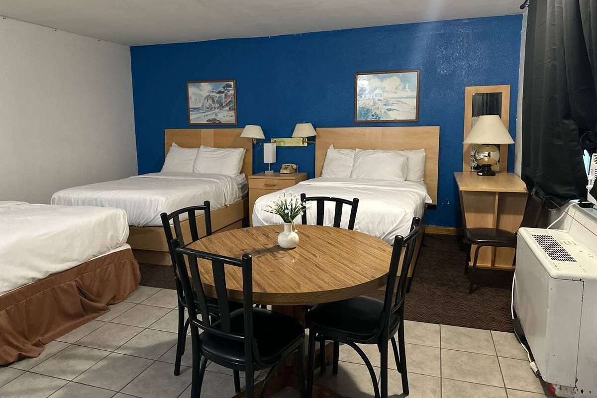 A hotel room with two single beds, a wooden table with four chairs, a small table with a lamp, and a heater.