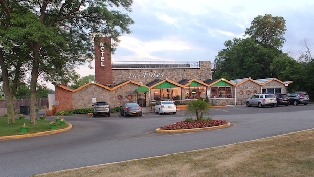 A motel with a brick facade, multiple windows, and a parking lot with several cars. - Tides Motor Inn at Stehli Beach (Locust Valley)