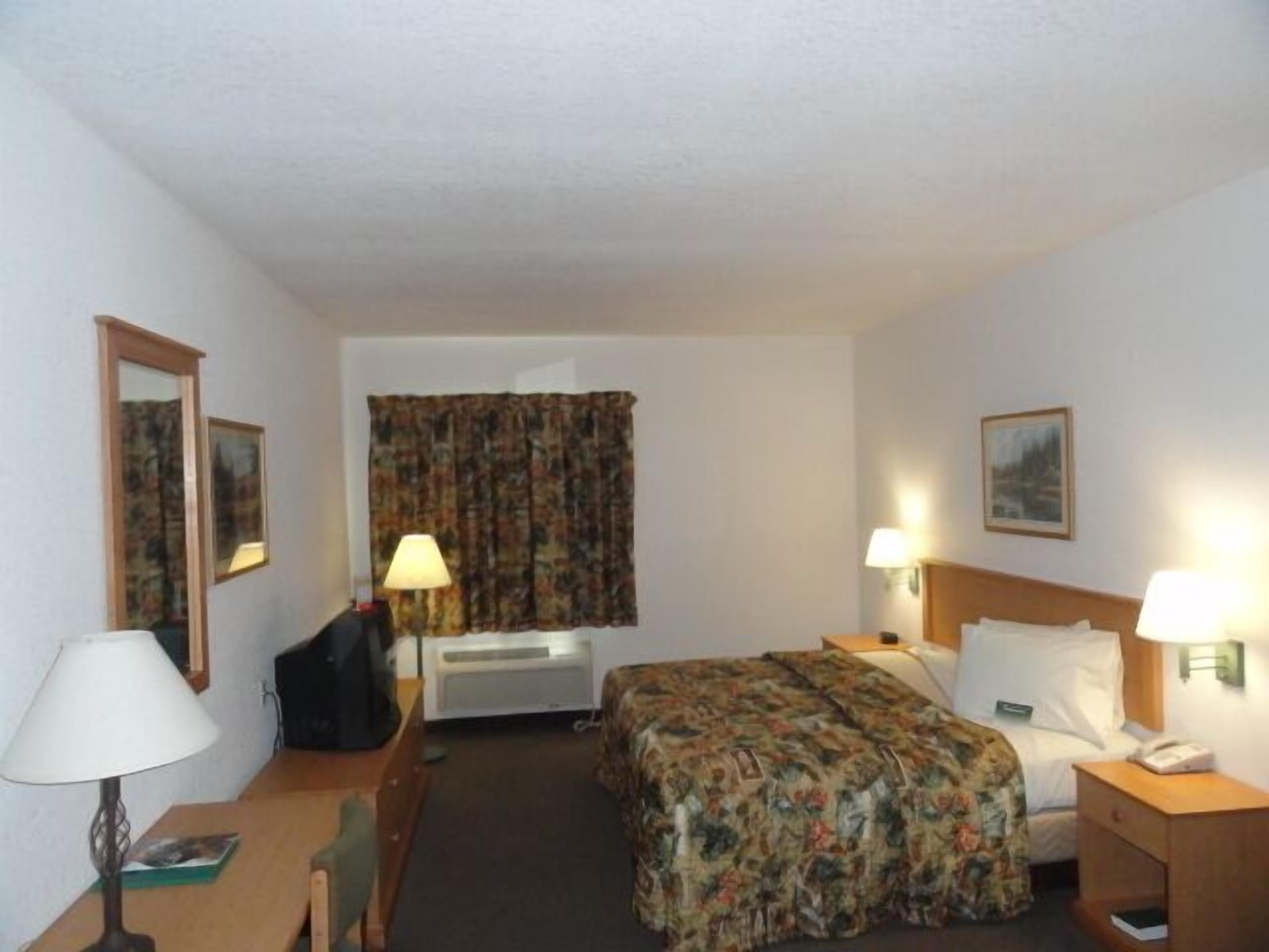A hotel room with a bed, a desk, a chair, a lamp, a bedside table, a telephone, and a window with curtains.