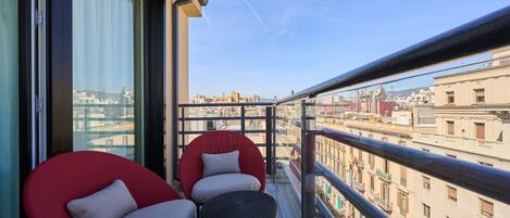 A balcony with two red chairs and a wicker ottoman, overlooking a cityscape with buildings and a clear sky.