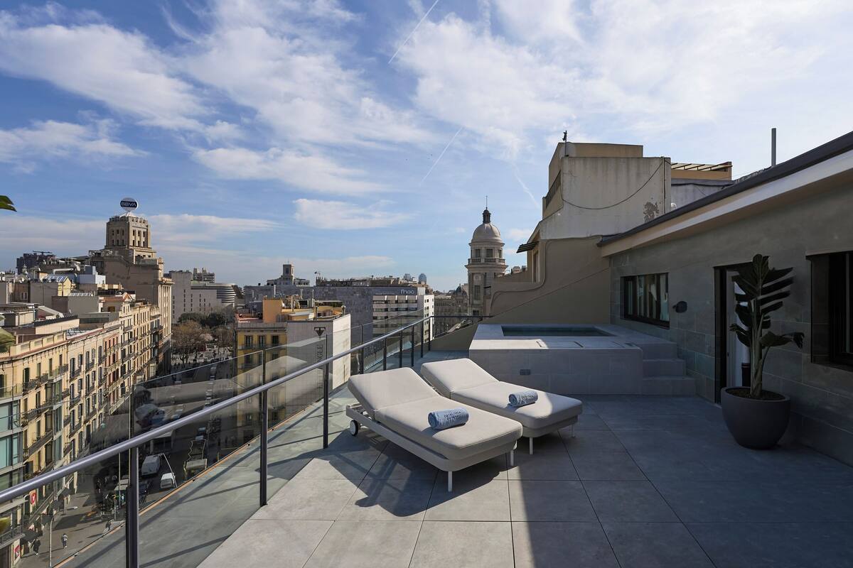 A rooftop terrace with lounge chairs and a hot tub, overlooking a cityscape with historic buildings.