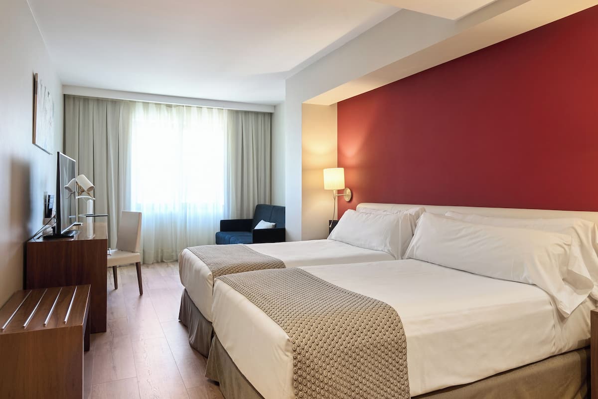 A hotel room with two beds, a desk, a chair, a TV, and a window with curtains.