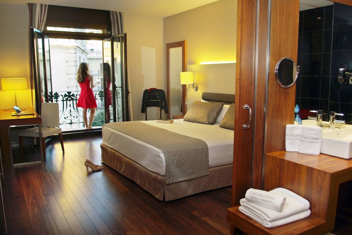 A hotel room with a bed, a desk, a mirror, and a bathroom with a sink and towel rack.