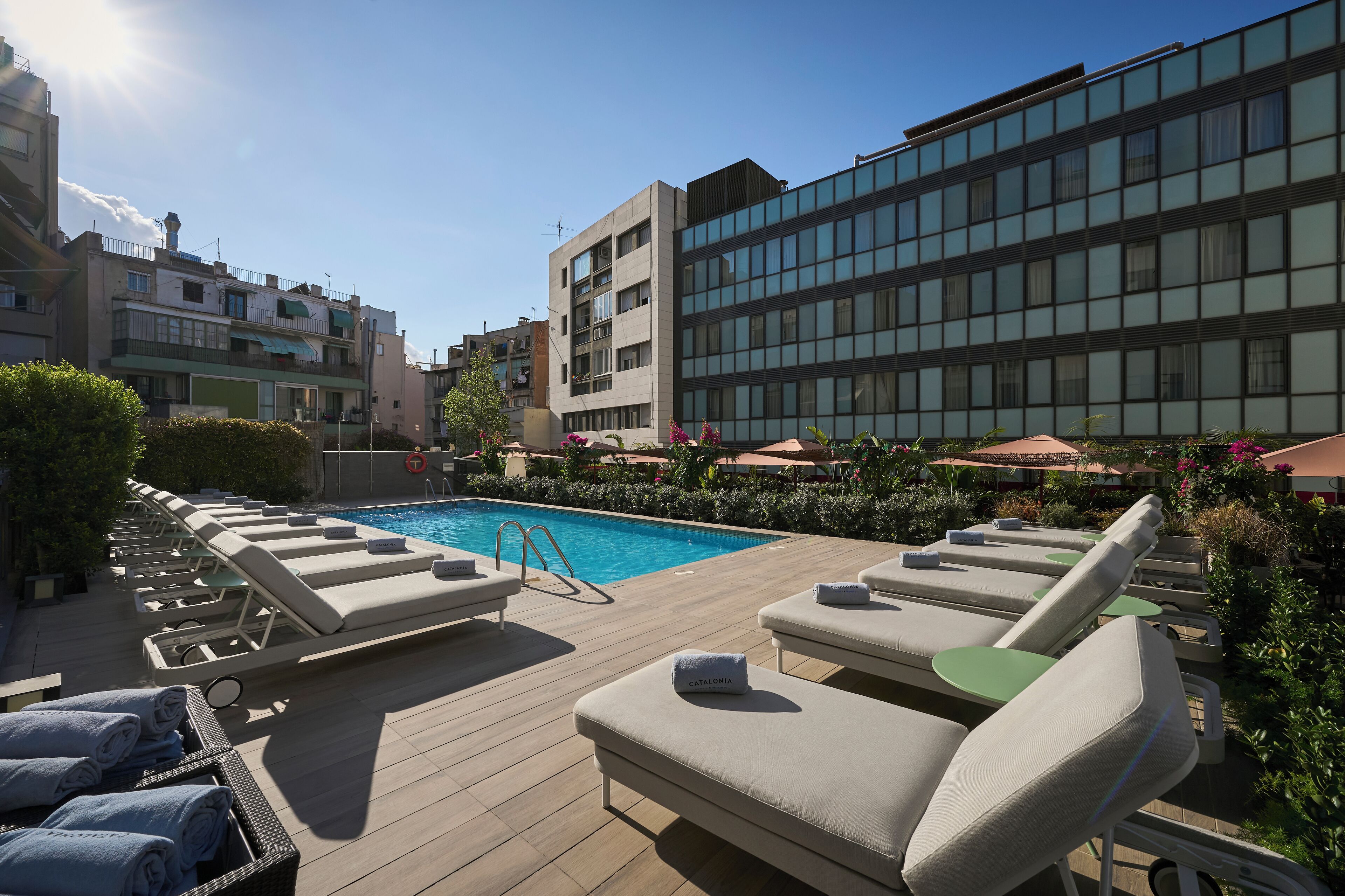 A rooftop pool area with lounge chairs and umbrellas, surrounded by greenery and modern buildings.