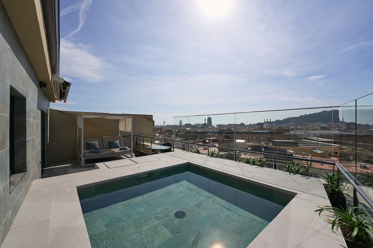 A rooftop pool with a city view, a seating area with cushions, and a glass railing.