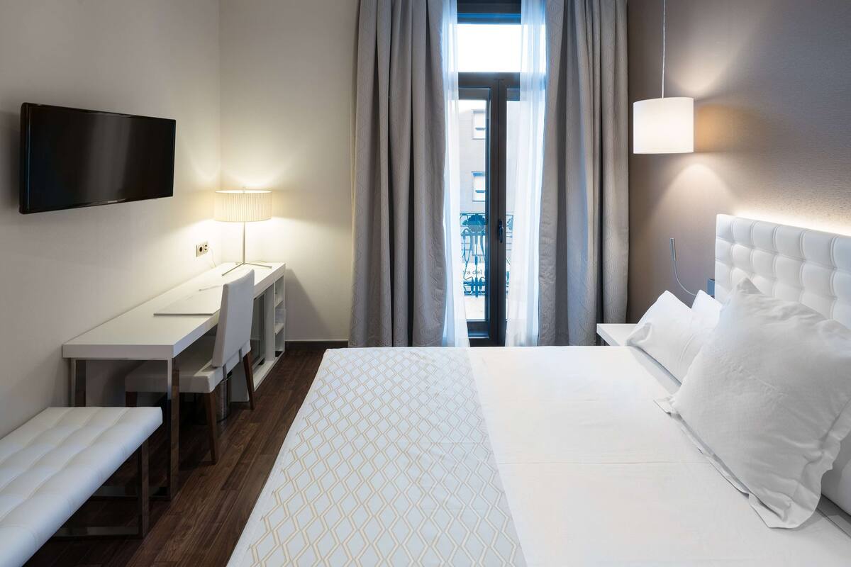 A hotel room with a bed, a desk, a chair, a TV, and a window with curtains.