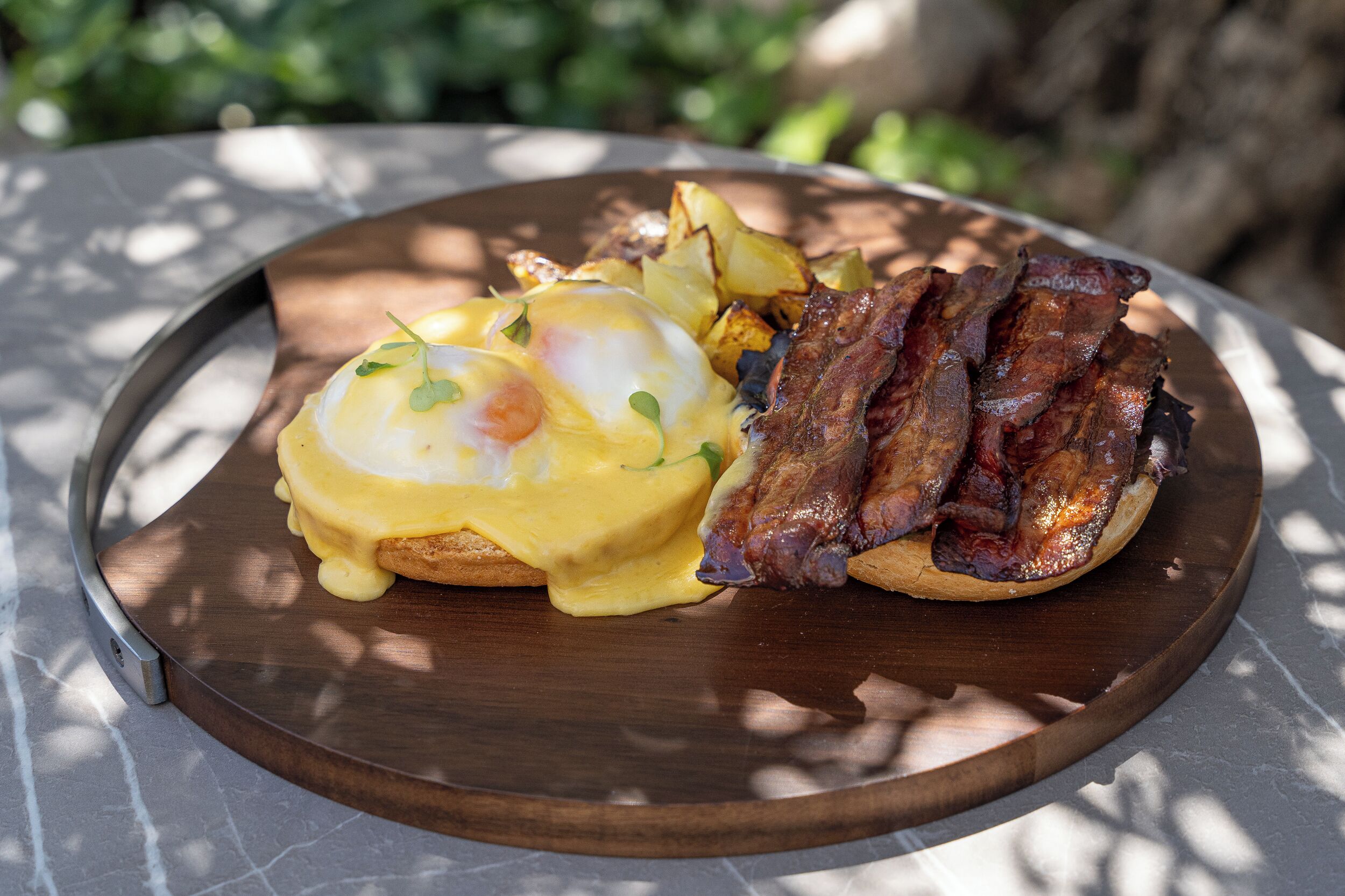 A breakfast plate with eggs Benedict, bacon, and potatoes on a wooden board.