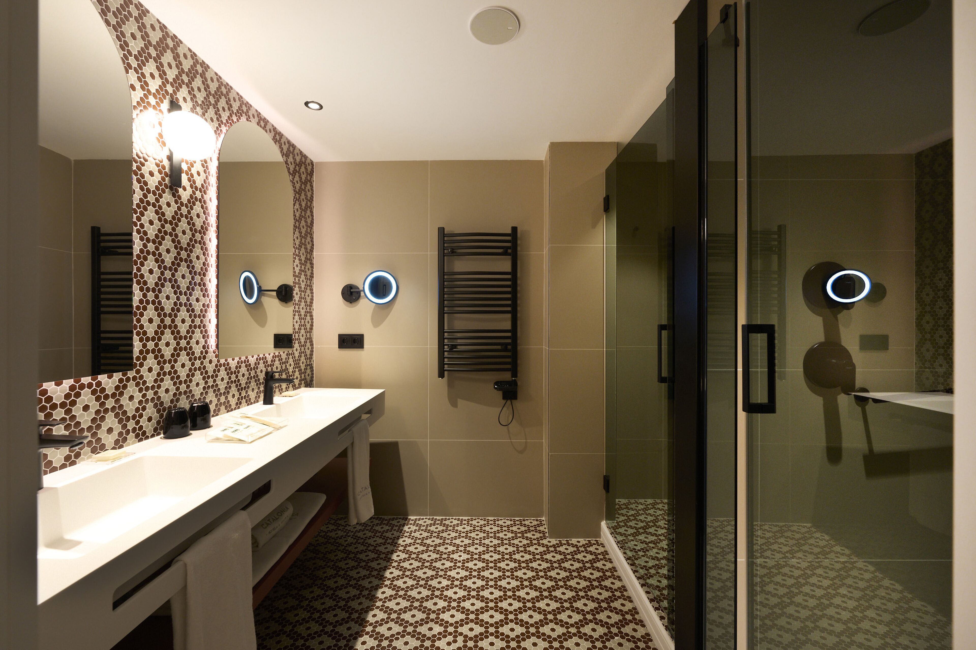 A modern bathroom with a large mirror, two sinks, a glass shower enclosure, and a patterned floor.