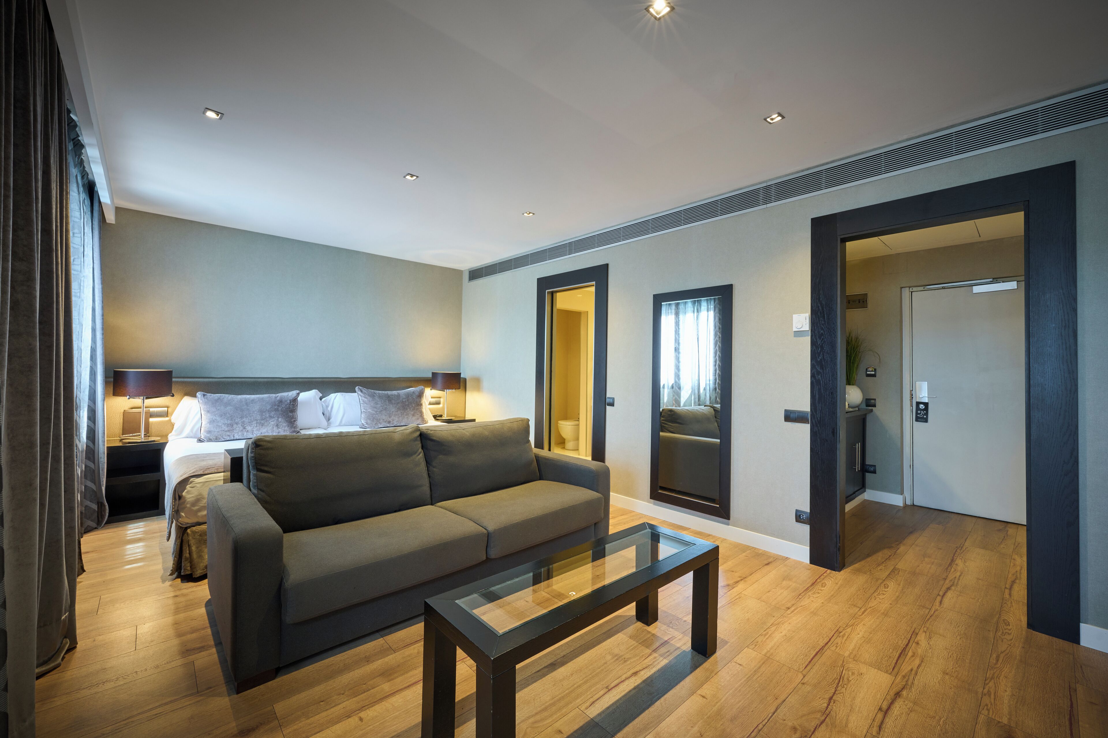 Una habitación de hotel moderna con sofá, una mesa de centro de vidrio, una cama con mesita de noche y un baño visible a través de una puerta abierta.