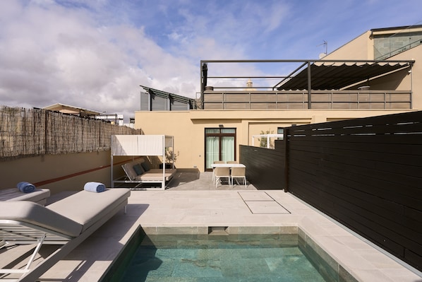 Uma área de piscina moderna ao ar livre, com espreguiçadeiras e uma mesa, cercada por um muro de madeira e um edifício com varanda.
