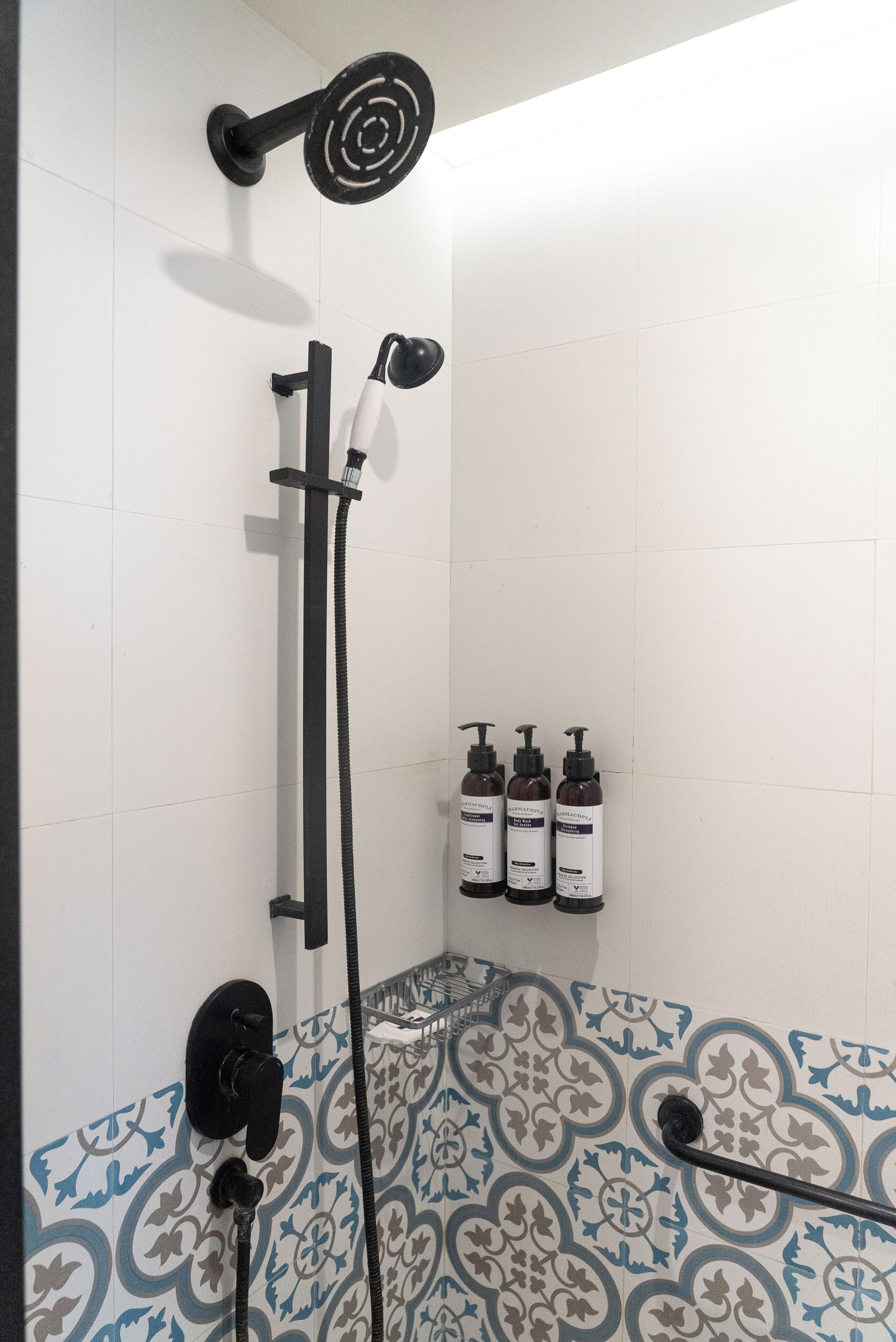 A shower area with a black handheld showerhead, a wall-mounted showerhead, and a handheld showerhead on a stand. There are three bottles of shampoo and conditioner on the wall.