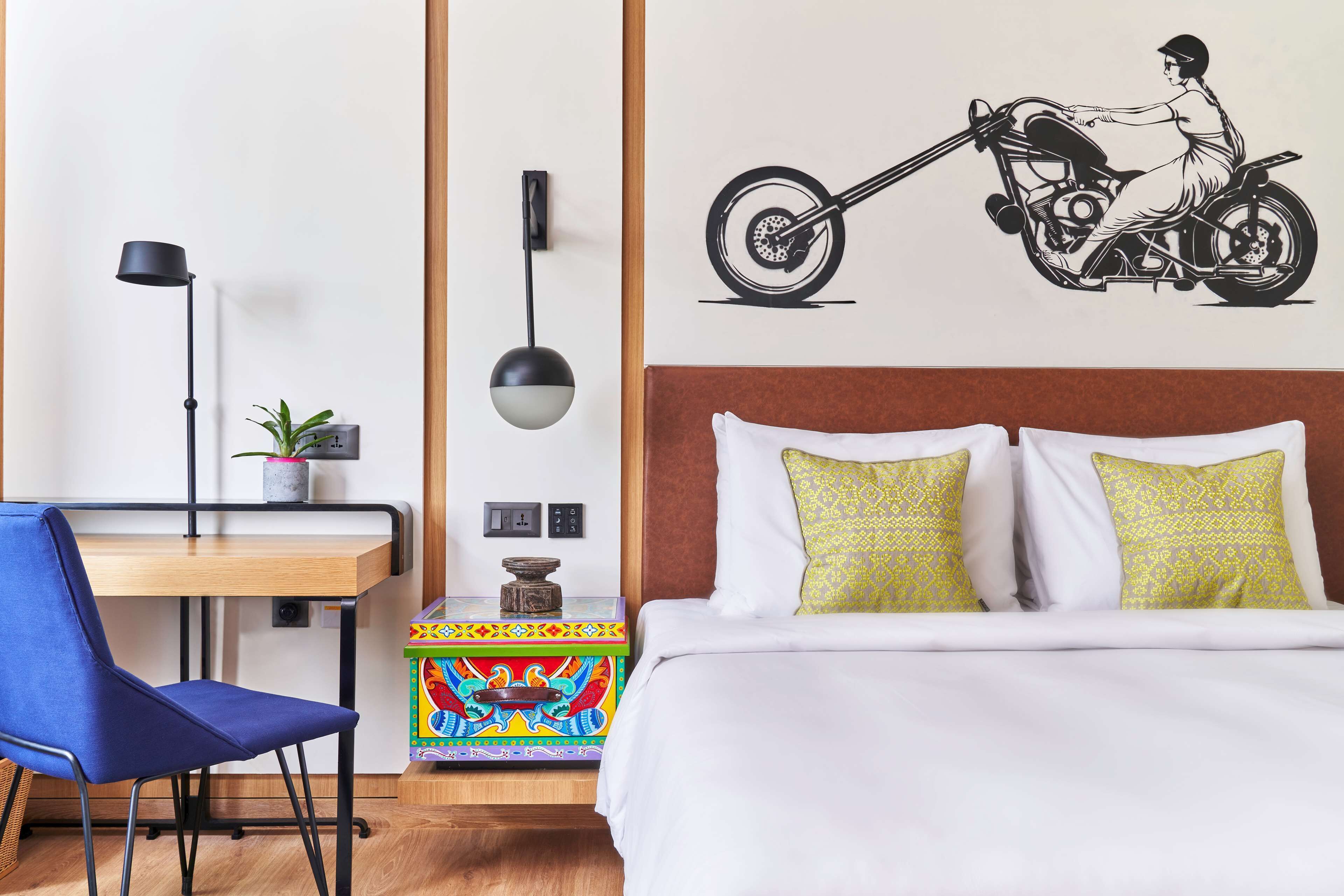 Una habitación de hotel con cama, escritorio, silla, lámpara y un adhesivo mural de una motocicleta.