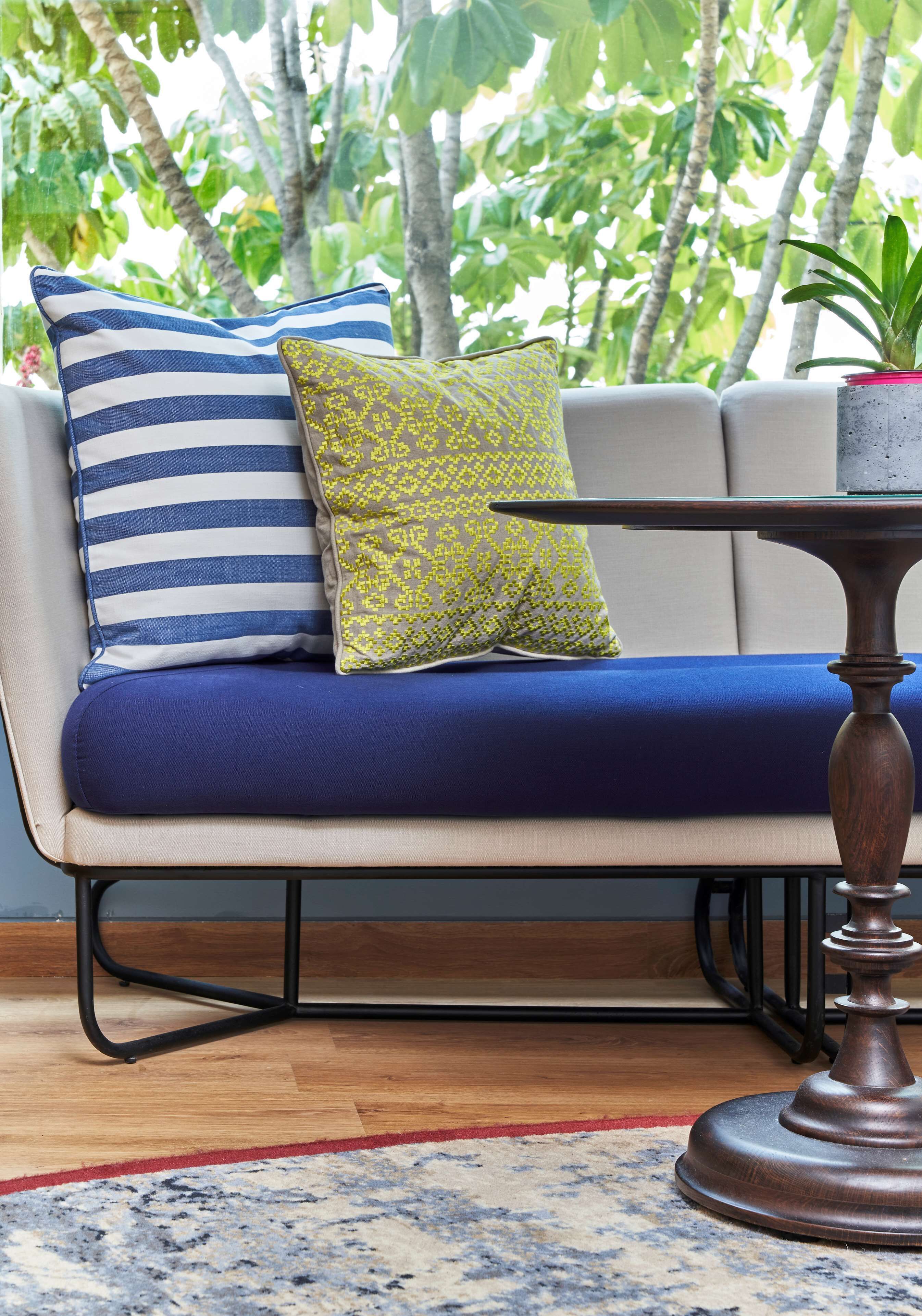 Ein modernes Sofa mit blauen Kissen und gemusterten Kopfkissen, ein runder Holztisch und Blick auf üppige, grüne Bäume.