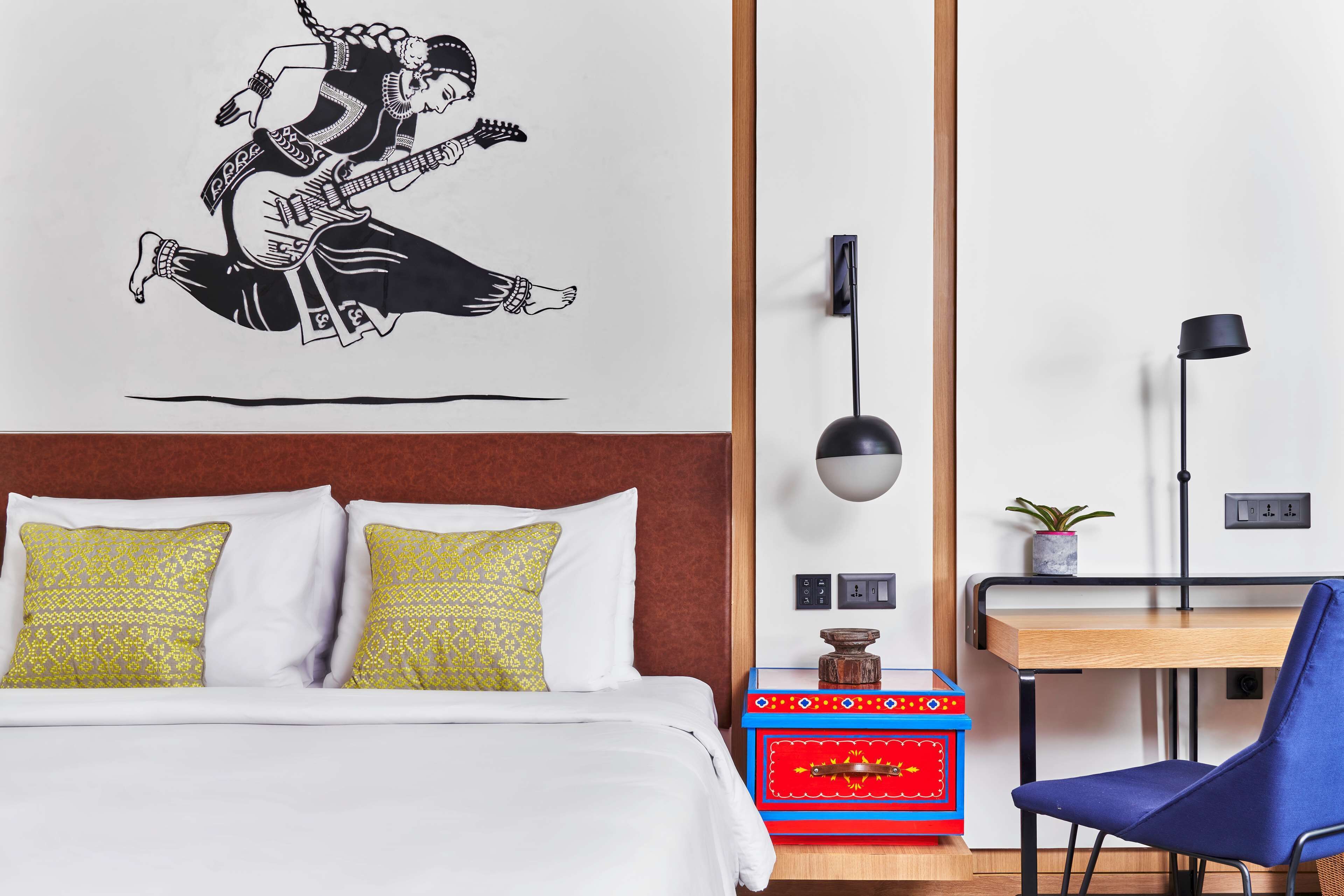 A hotel room with a bed, a desk, a chair, a wall-mounted lamp, and a decorative wall piece.