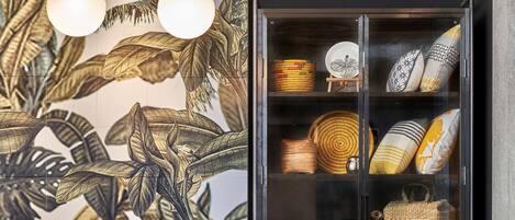 Una moderna zona de recepción con un mostrador de hormigón, un mural de hojas tropicales y una vitrina de cristal con cestas tejidas y cojines decorativos.