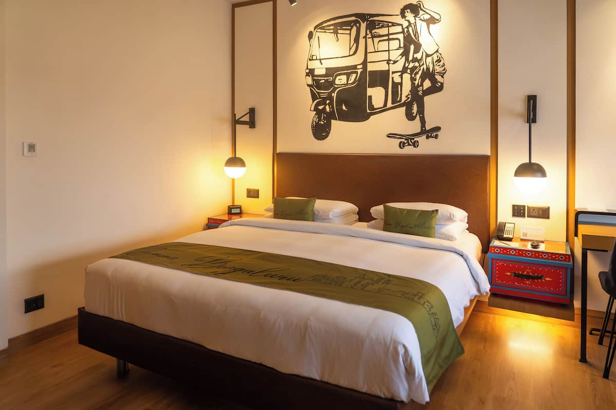 Un lit bien fait, recouvert d’une couverture verte, un luminaire fixé au mur et une œuvre d’art représentant une personne sur une planche à roulettes.