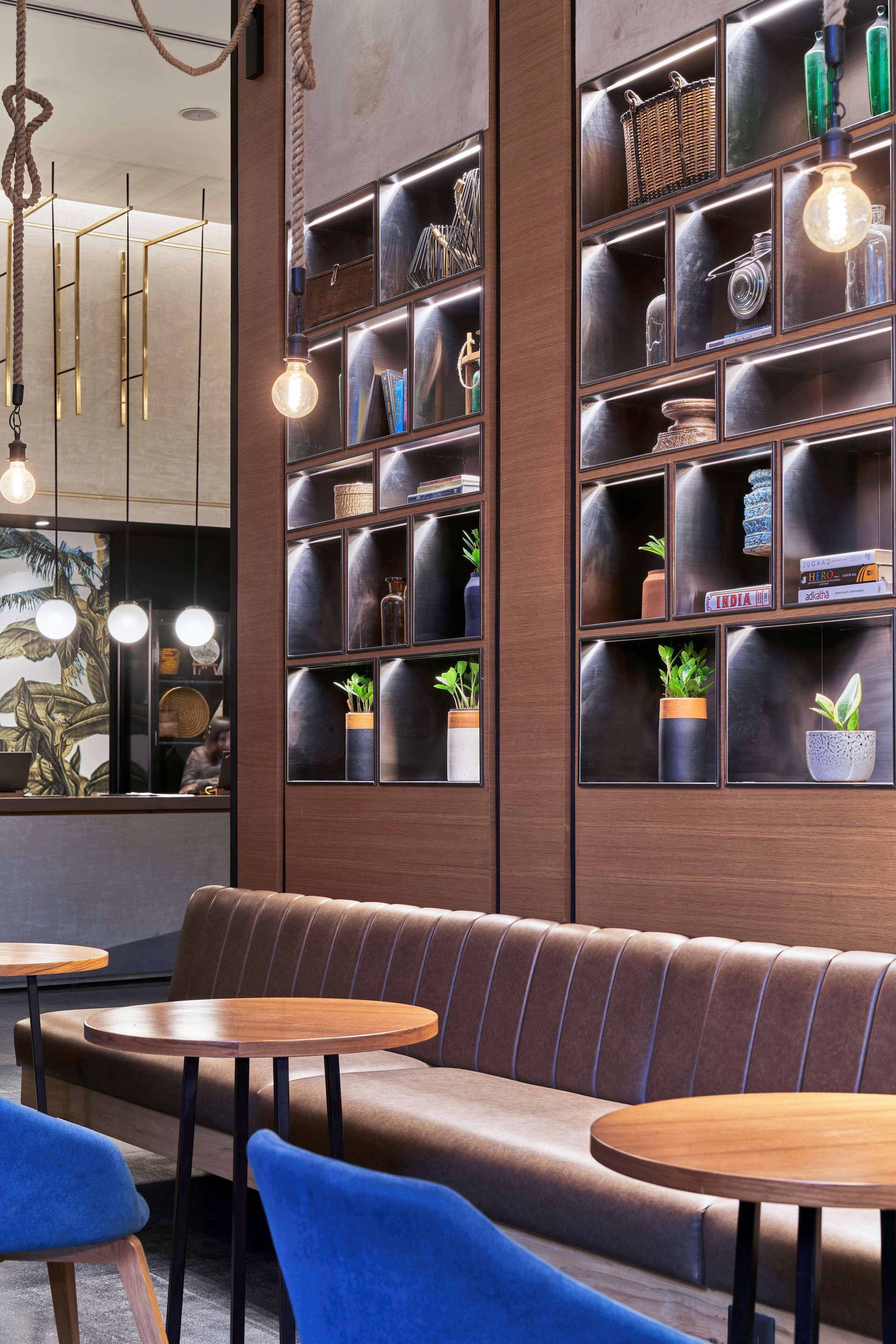 A modern interior with a wooden bench and round tables, a wall-mounted shelf with decorative items, and hanging light fixtures.