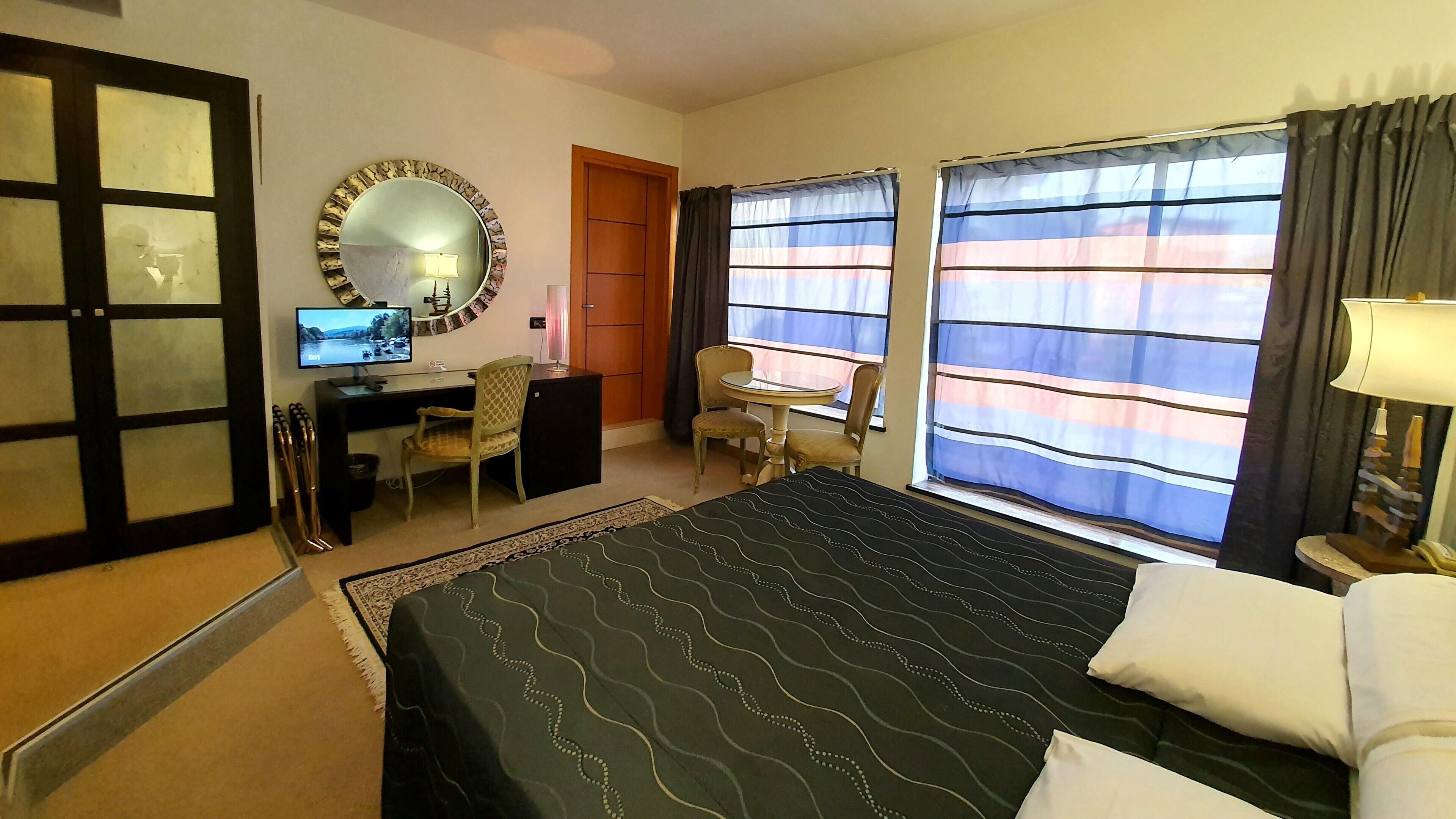 A hotel room with a bed, desk, chair, TV, and large windows with curtains.