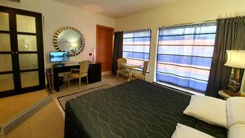 A hotel room with a bed, desk, chair, TV, and large windows with curtains.
