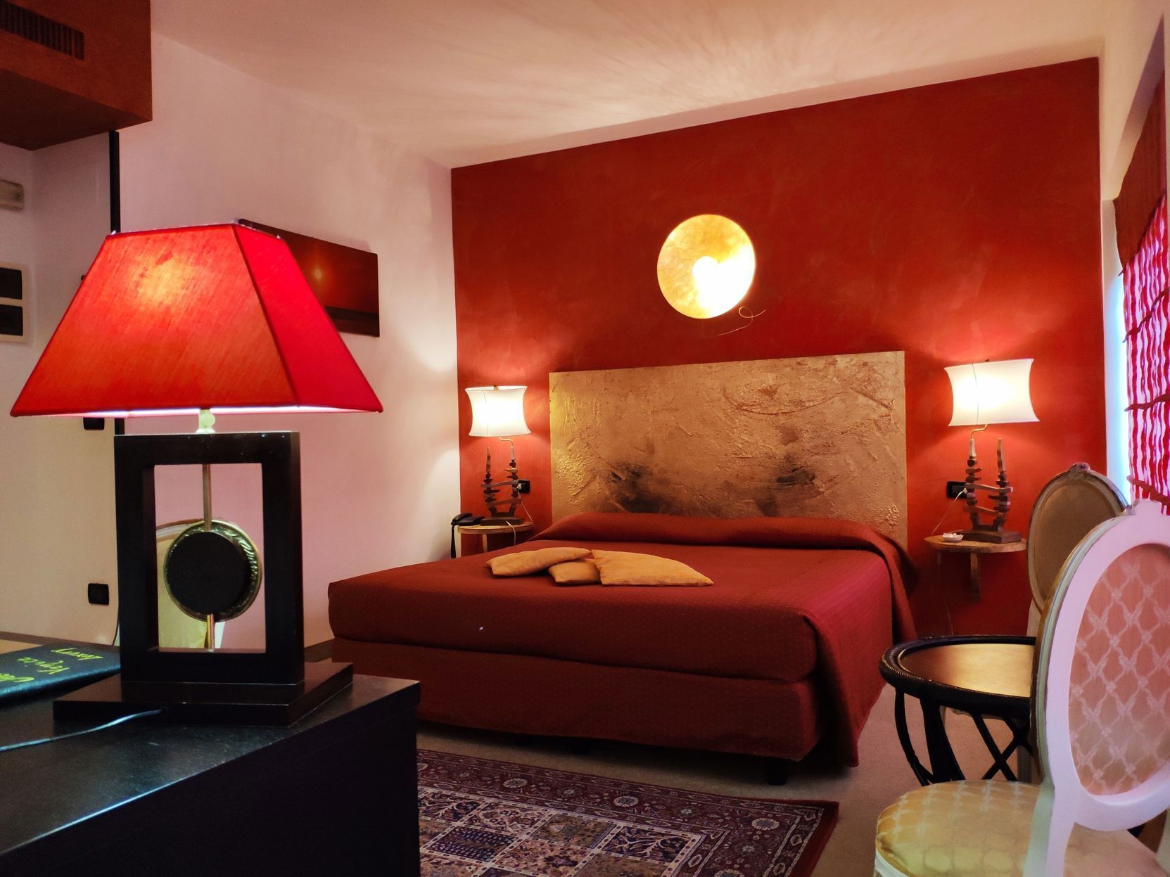 Een slaapkamer met een rood bed, twee bedlampen, een ronde decoratie aan de muur, een klein rond tafeltje en een stoel.