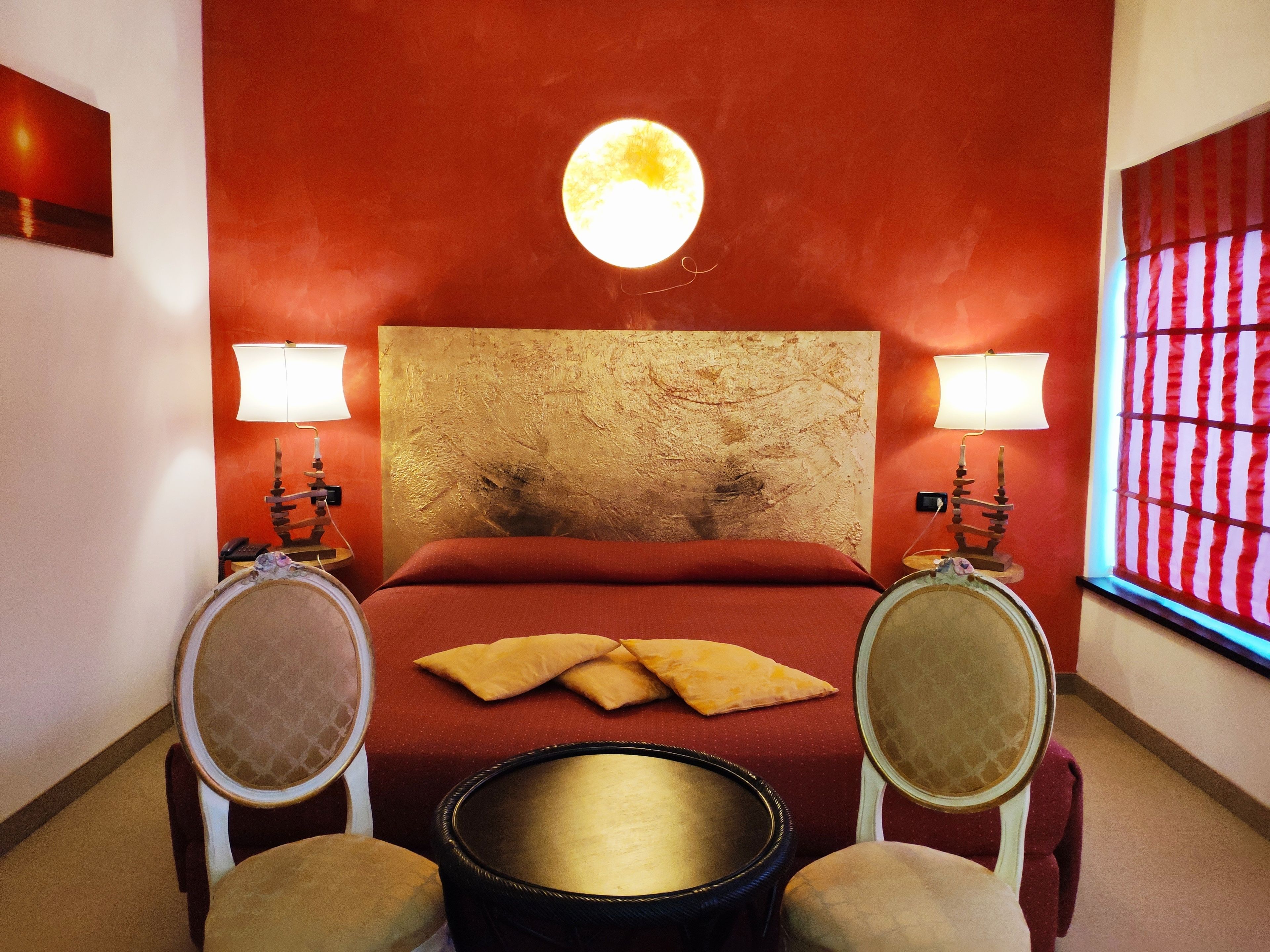 Habitación de hotel con una pared roja, un espejo redondo, una cama con ropa de cama roja, dos lámparas de noche y una mesa redonda con una silla.