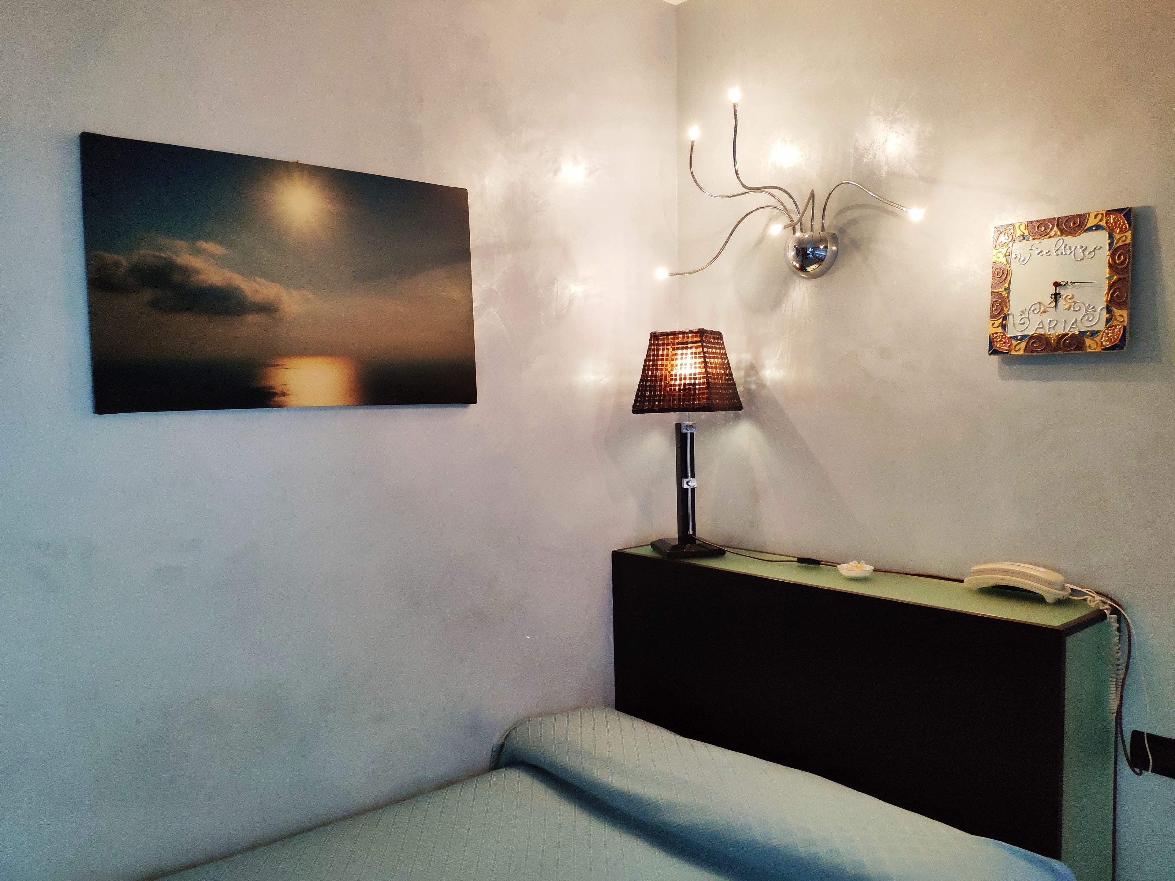 Ein Schlafzimmer mit einem Bett, einem Nachttisch mit Lampe, einem Wandtelefon, einem gerahmten Bild und einer Wandleuchte.