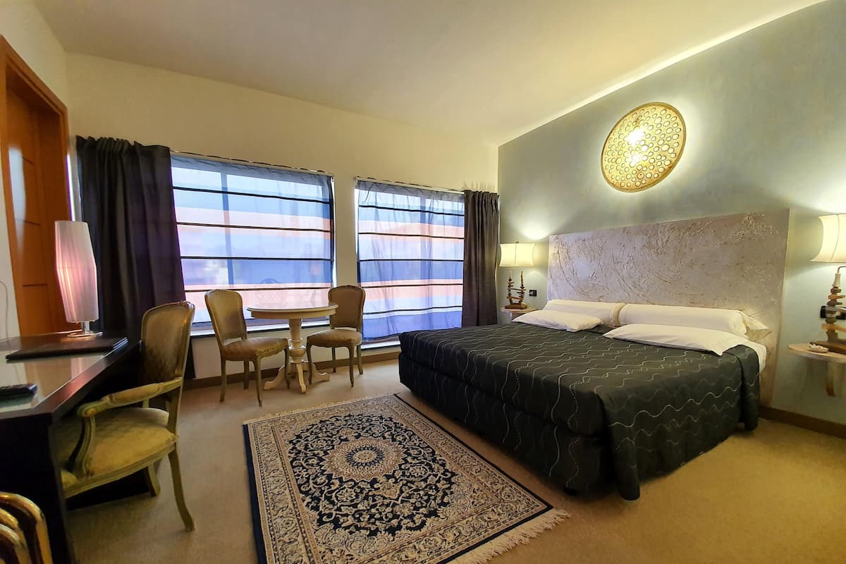 Ein Hotelzimmer mit einem Bett, einem Schreibtisch mit Lampe, zwei Stühlen, einem Tisch, einem Teppich und großen Fenstern mit Vorhängen.