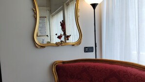 Ein Zimmer mit einem roten, gepolsterten Sofa, einem goldfarbenen Spiegel, einer Lampe und einem Fenster mit weißen Vorhängen.