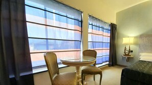 Ein Hotelzimmer mit einem runden Glastisch, zwei Stühlen, einem Bett, einem Nachttisch mit Lampe und großen Fenstern mit gestreifteren Vorhängen.