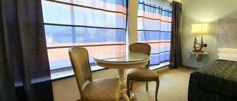 Habitación de hotel con una mesa redonda de cristal, dos sillas, una cama, una mesita de noche con lámpara y ventanas grandes con cortinas a rayas.