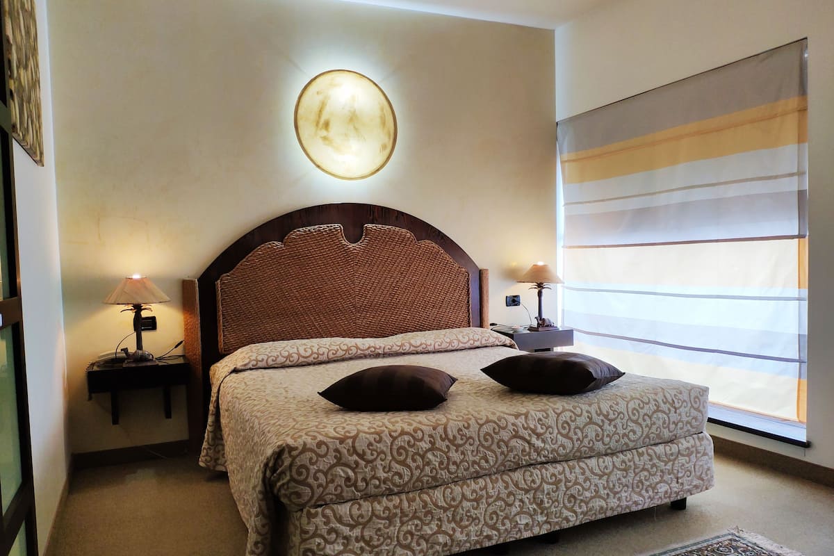 Ein Schlafzimmer mit einem großen Bett, zwei Nachttischlampen, einer Wandleuchte und einem Fenster mit Jalousien.