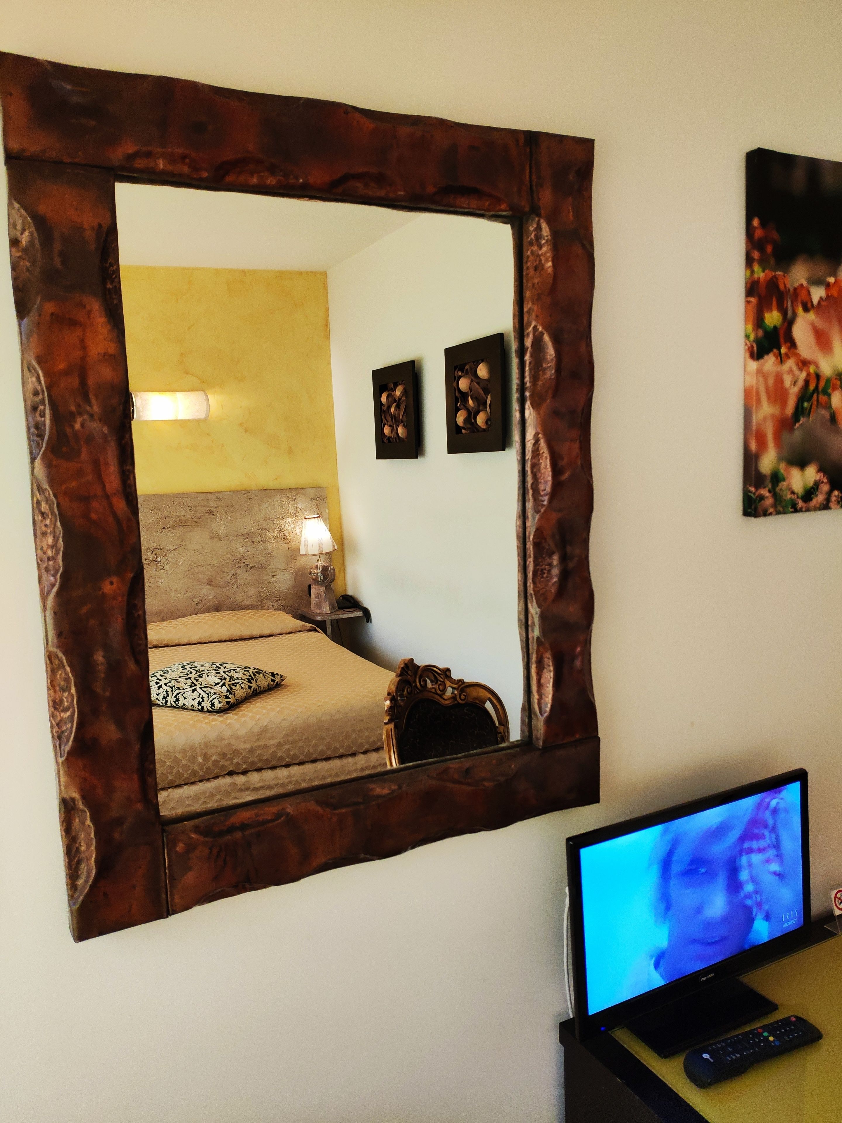 A bedroom with a large mirror, a bed with a patterned pillow, a bedside lamp, and a television on a stand.