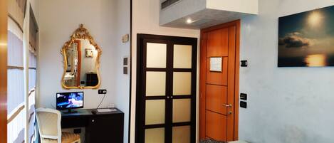 A hotel room with a bed, a desk with a computer, a chair, a mirror, and a door.