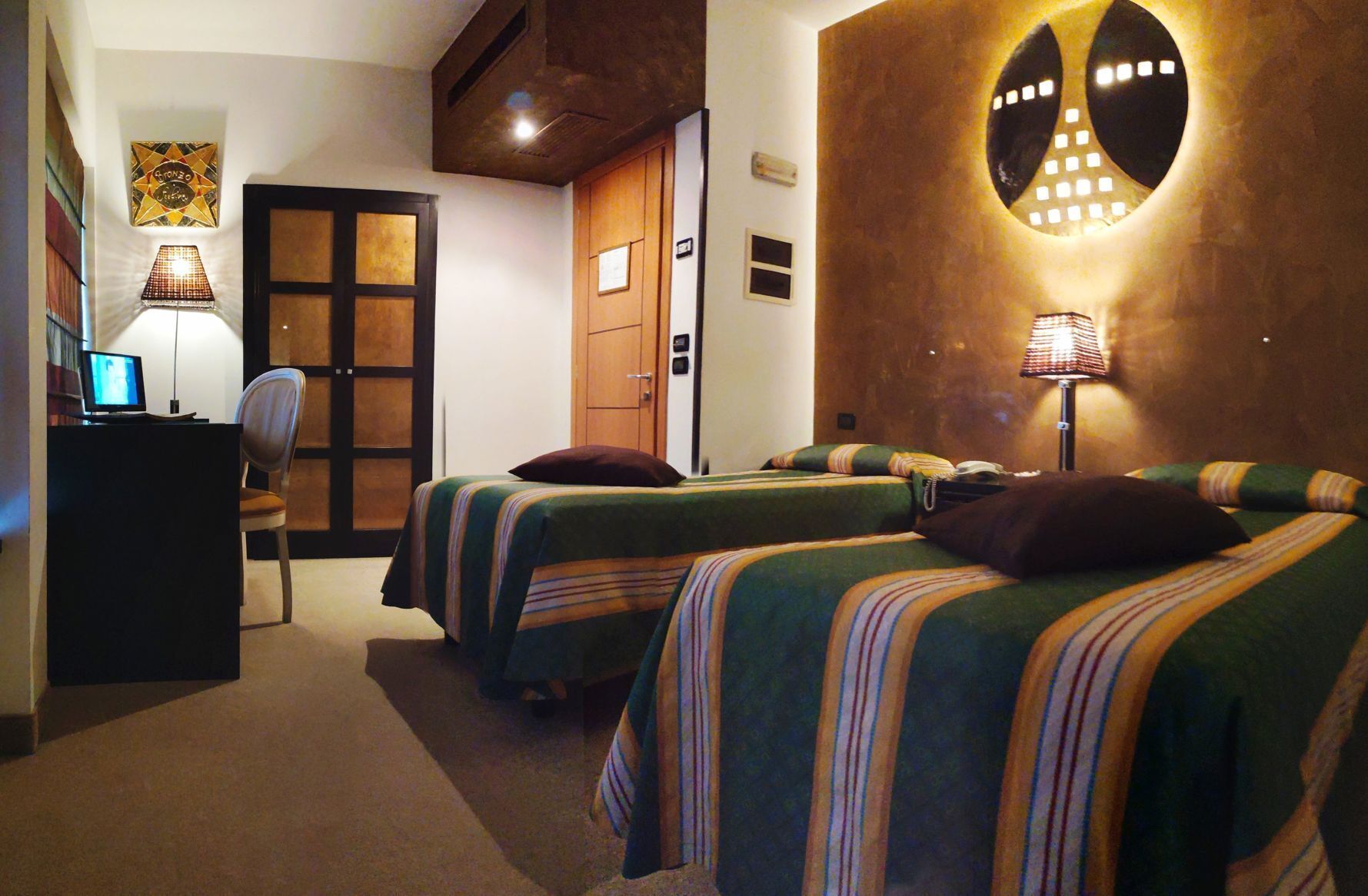 A hotel room with two beds, a desk with a computer, a chair, a door, and a wall-mounted lamp.