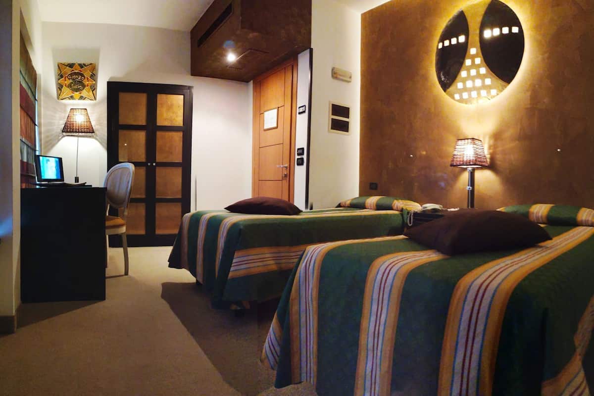 Ein Hotelzimmer mit zwei Betten, einem Schreibtisch mit Computer, einem Sessel, einer Tür und einer Wandlampe.