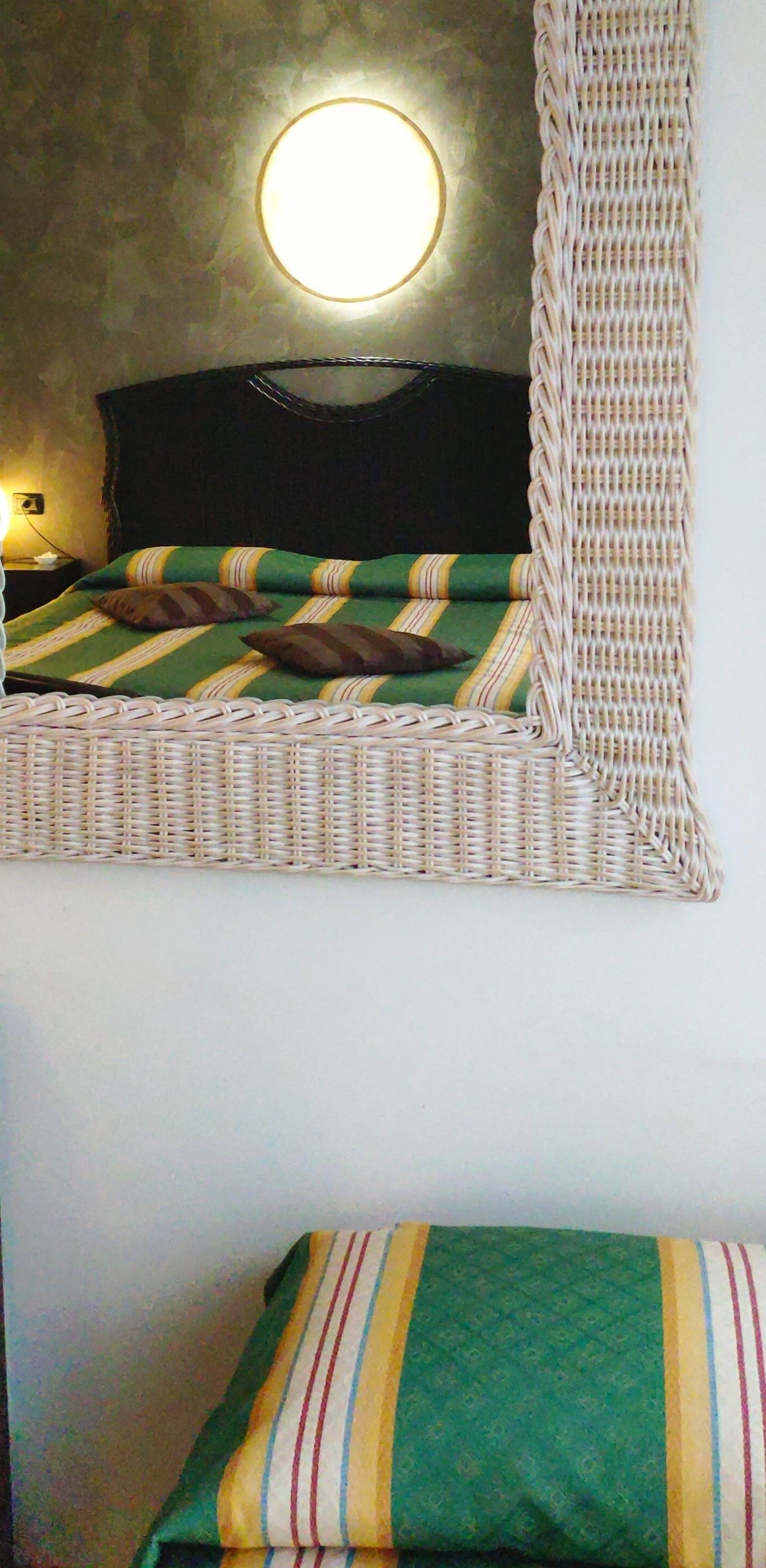 Ein Bett mit einer grün-gelb gestreiften Tagesdecke, einer runden Wandleuchte und einem Korbstuhl.