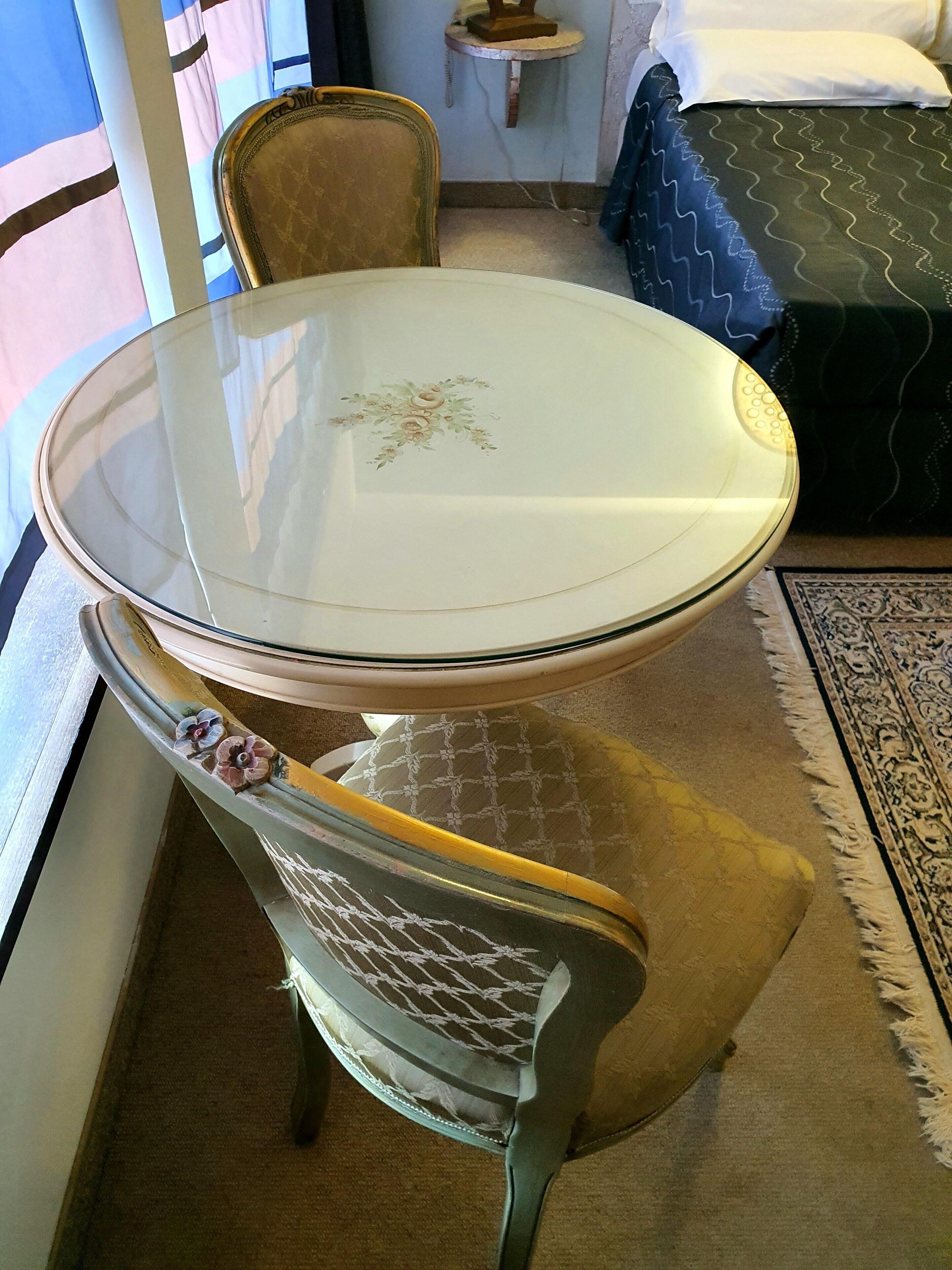 A room with a glass-top table, a floral-patterned chair, a bed with a blue patterned bedspread, and a patterned rug.
