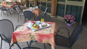 Une table dressée avec des aliments pour le petit-déjeuner, notamment des fruits, des viennoiseries et une tasse de café, entourée de chaises dans un espace de restauration en plein air.