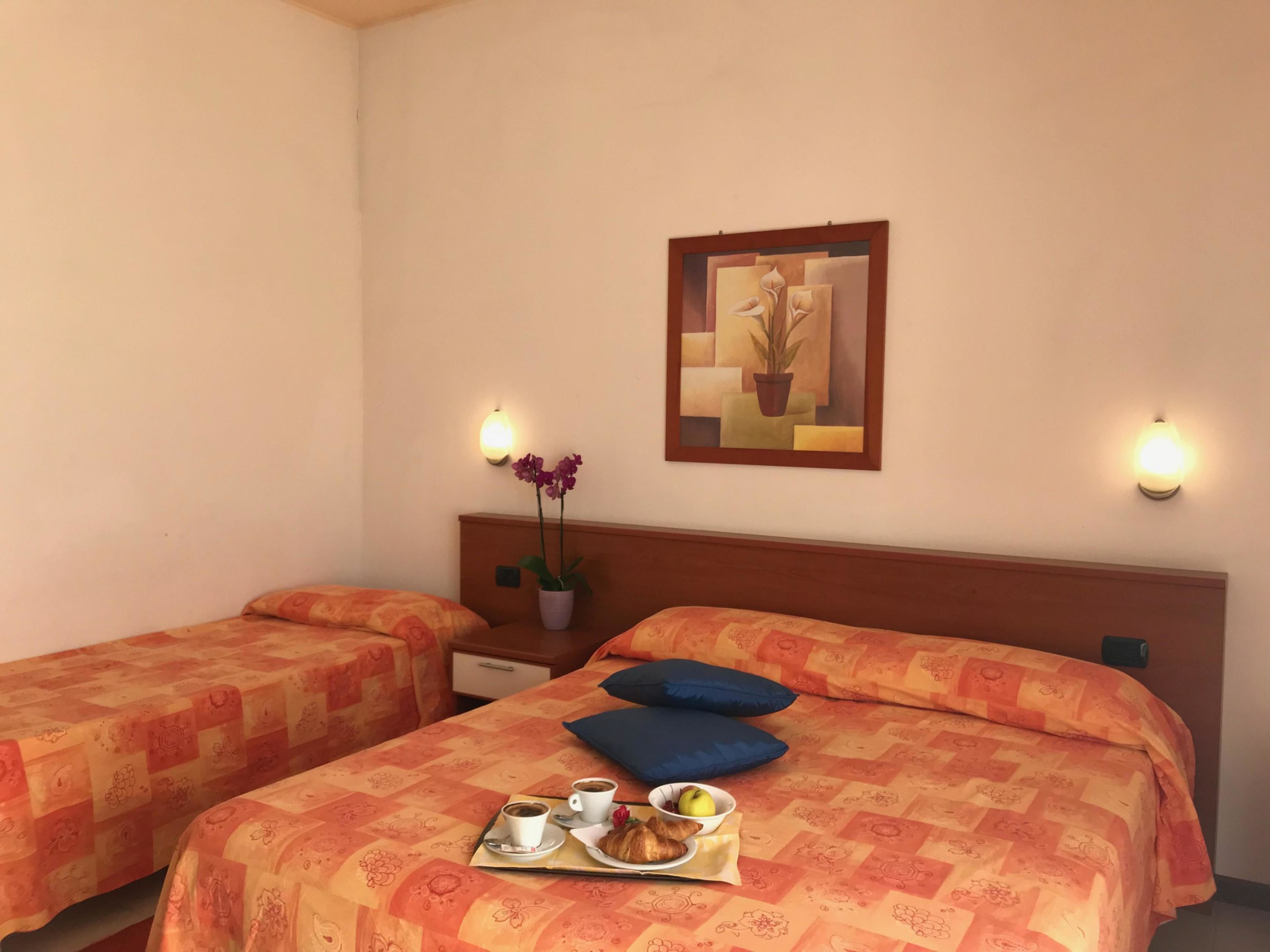 Een hotelkamer met twee bedden, elk met bijpassend oranje beddengoed. Op het bed staat een dienblad met een croissant, een citroen en een kop koffie. Op het nachtkastje staat een potplant, en aan de muur hangt een ingelijste afbeelding.