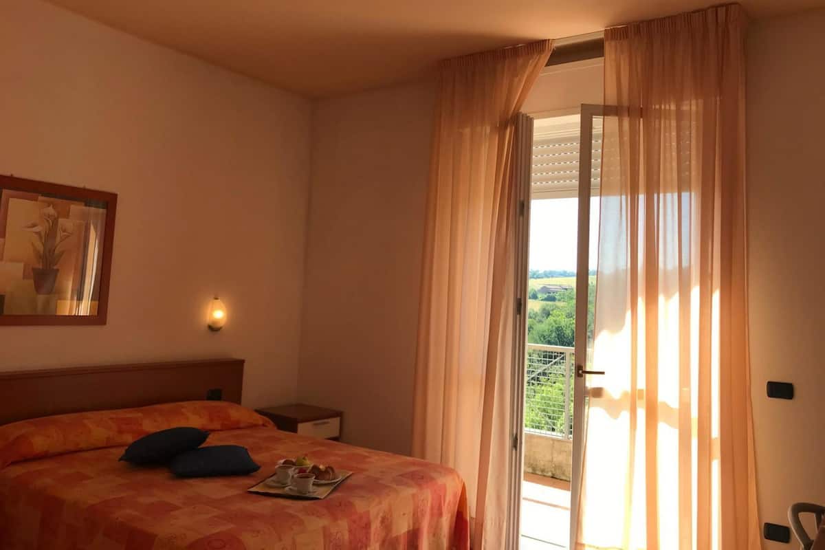 Une chambre d’hôtel avec un lit, une table de chevet, un tableau et un balcon avec des rideaux.