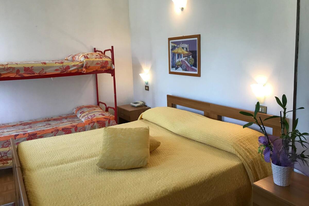 Une chambre avec un lit superposé, un lit simple, une table de chevet avec une lampe, un luminaire fixé au mur et un tableau encadré.