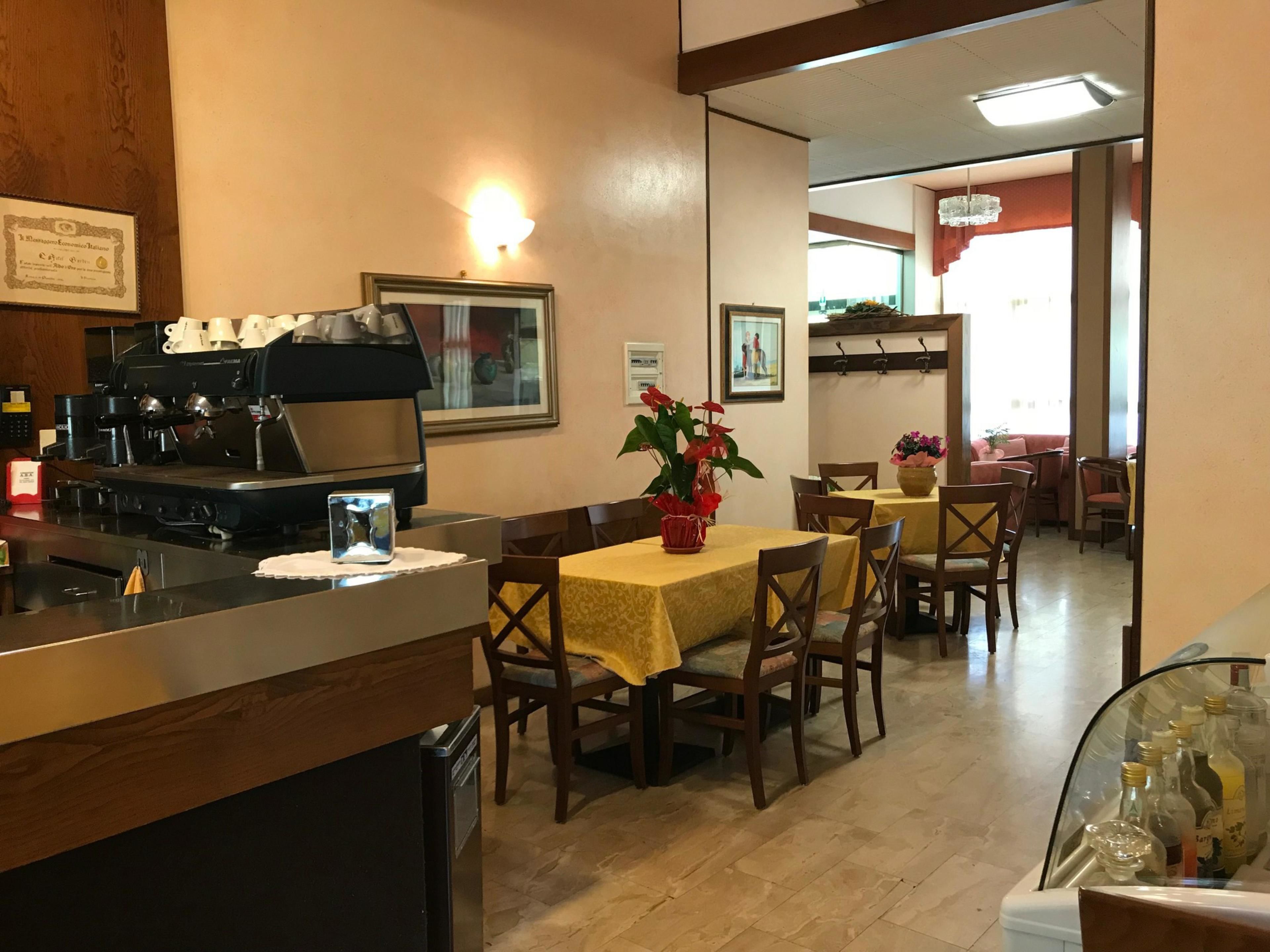 A restaurant with a coffee machine, tables with yellow tablecloths, and chairs. There are framed pictures on the wall and a window with a view of the outside.