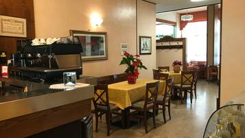 A restaurant with a coffee machine, tables with yellow tablecloths, and chairs. There are framed pictures on the wall and a window with a view of the outside.