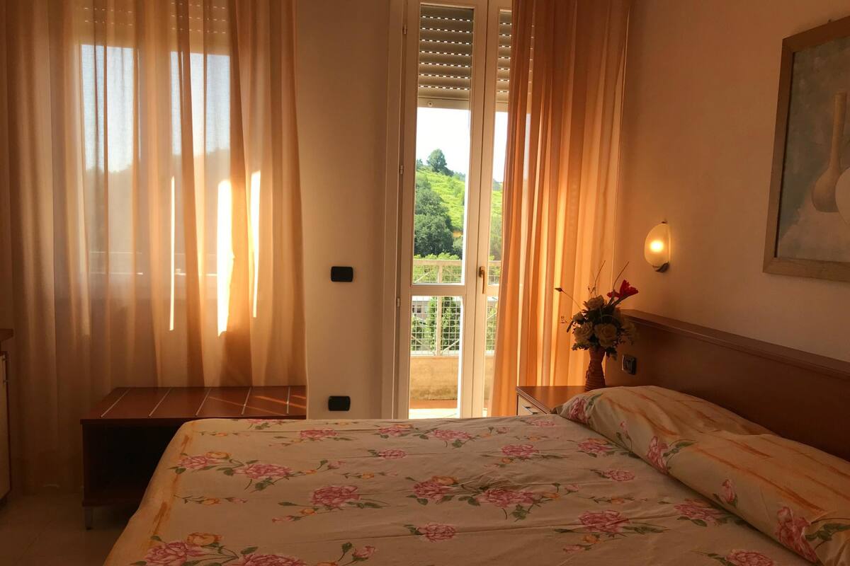 Une chambre à coucher avec un lit, des rideaux, une porte, une table de chevet et une lampe fixée au mur.