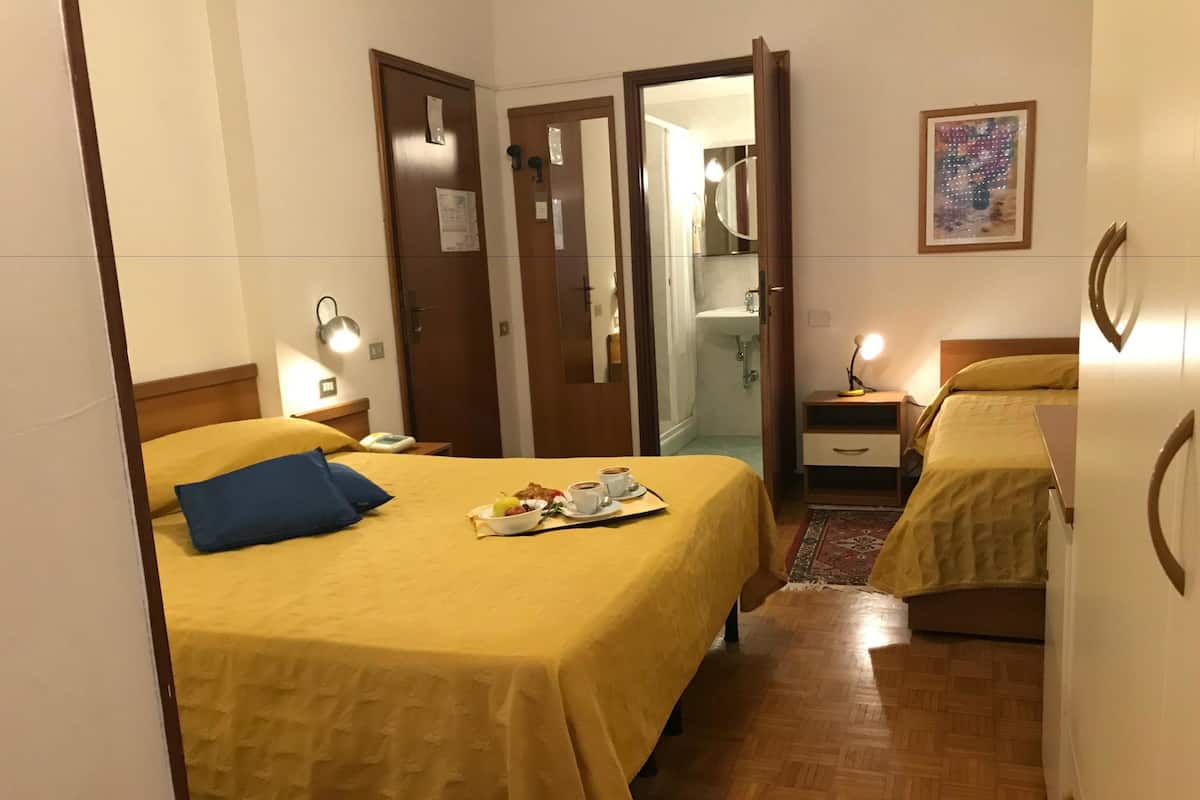 Une chambre d’hôtel avec deux lits, une salle de bain visible à travers une porte ouverte, une table de chevet avec une lampe et un petit tapis sur le sol.