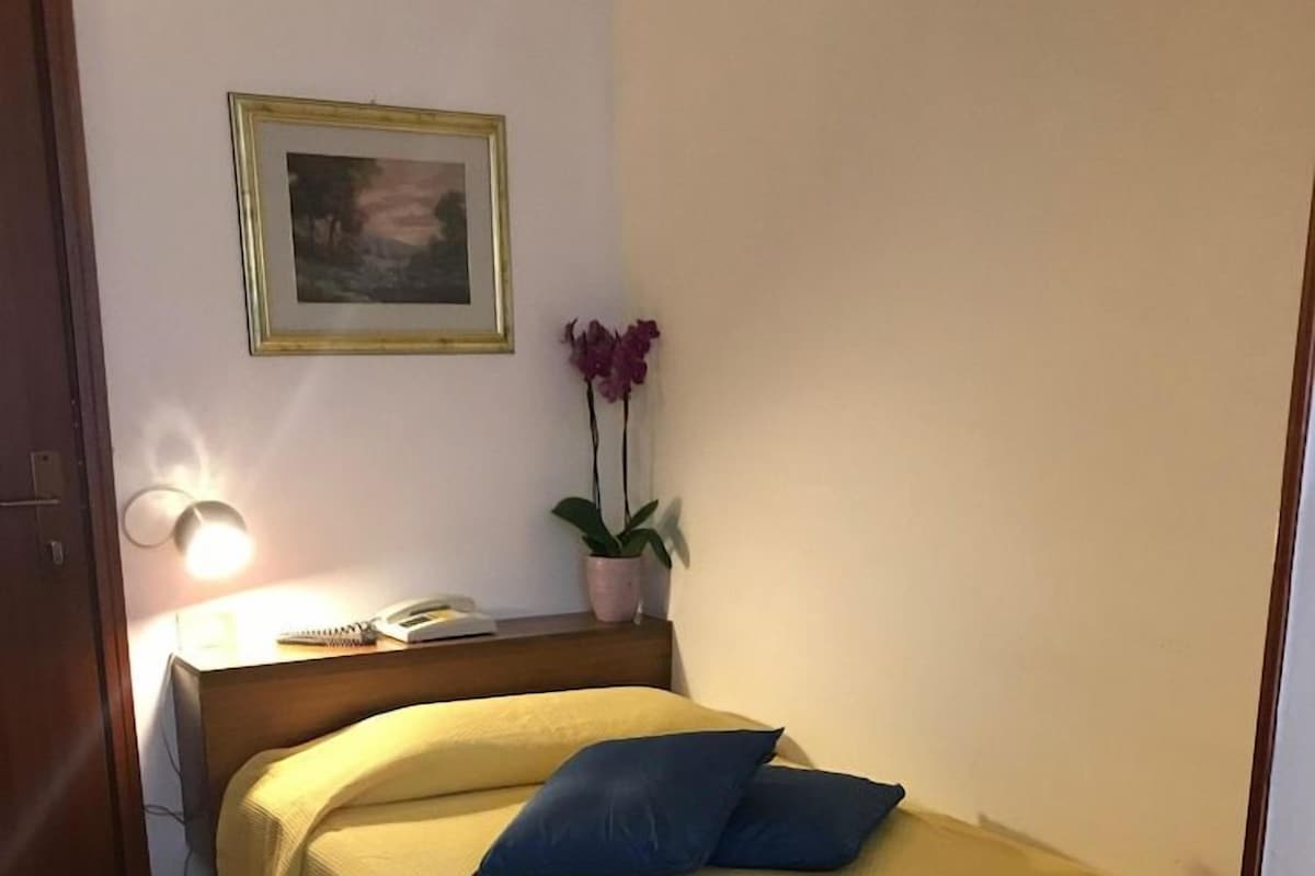 Une petite chambre à coucher, bien rangée, avec un lit, une table de chevet, une lampe et un tableau encadré au mur.