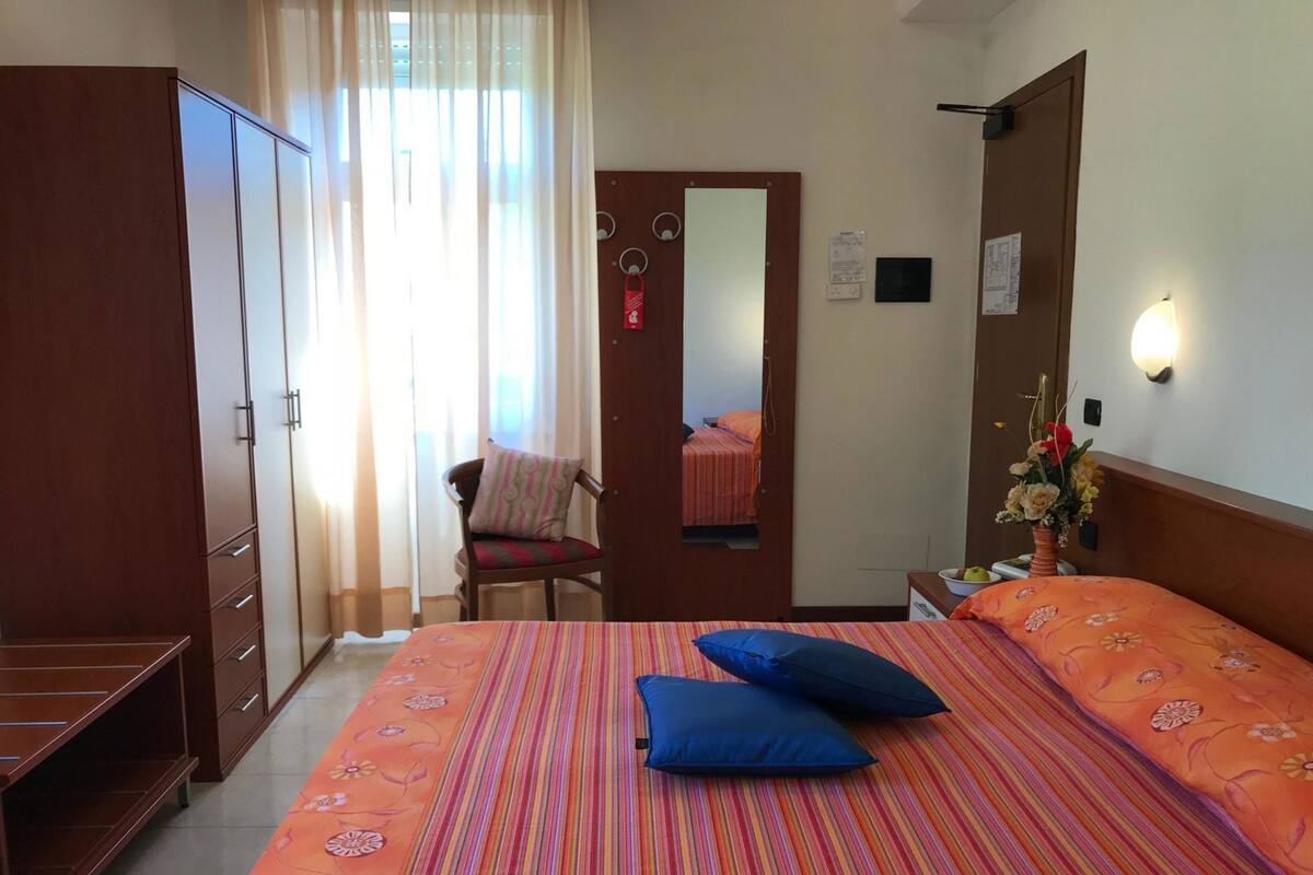 Une chambre d’hôtel comprenant un lit, une chaise, une armoire, un miroir et une table de chevet ornée d’un vase de fleurs.