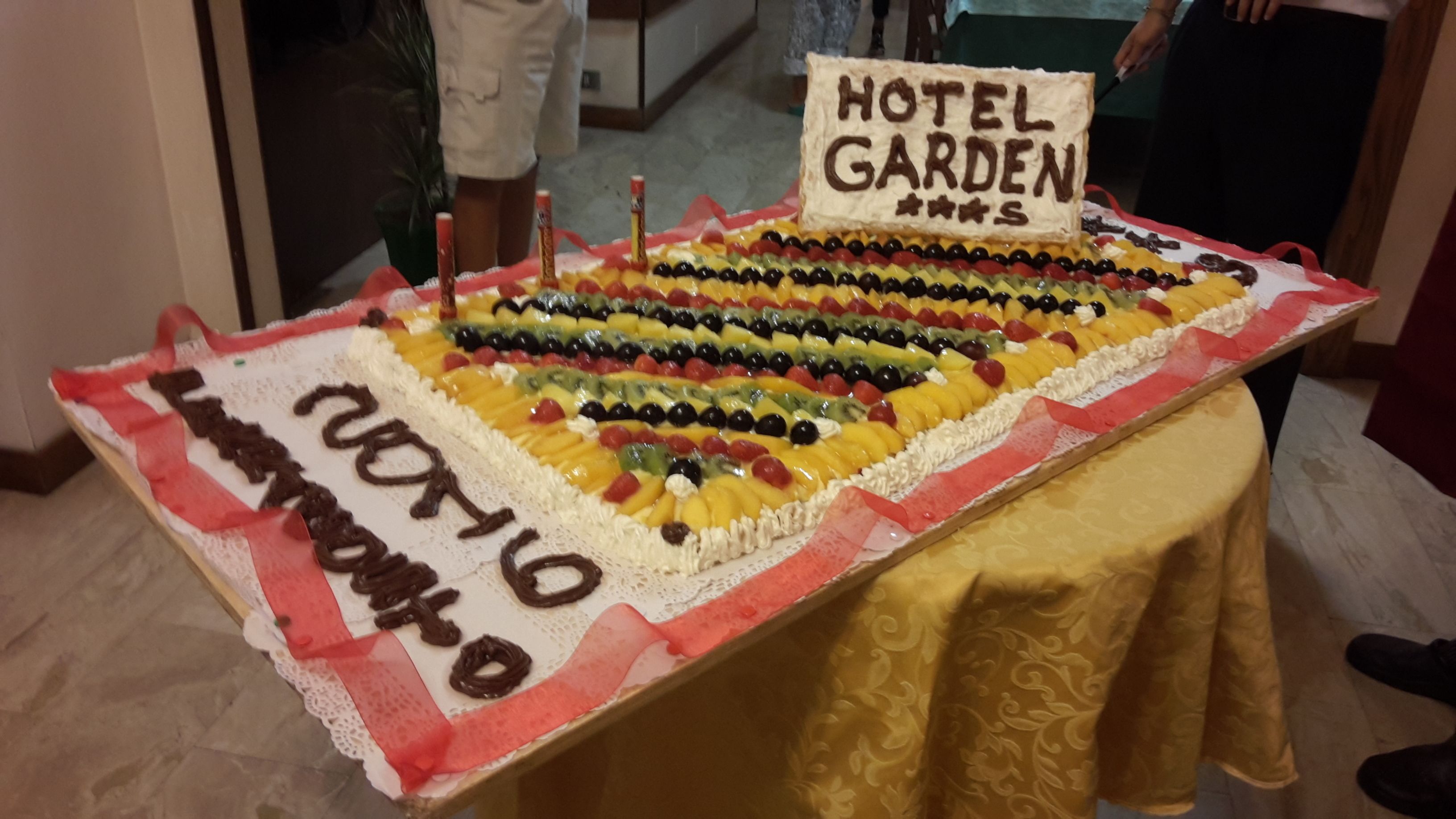 Uma mesa com um bolo grande coberto de frutas e um letreiro com a inscrição 'HOTEL GARDEN'.
