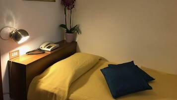 Ein ordentlich bezogenes Bett mit einer gelben Decke und einem blauen Kissen, eine Nachttischlampe, ein Telefon und ein Blumentopf auf einem Holznachttisch.