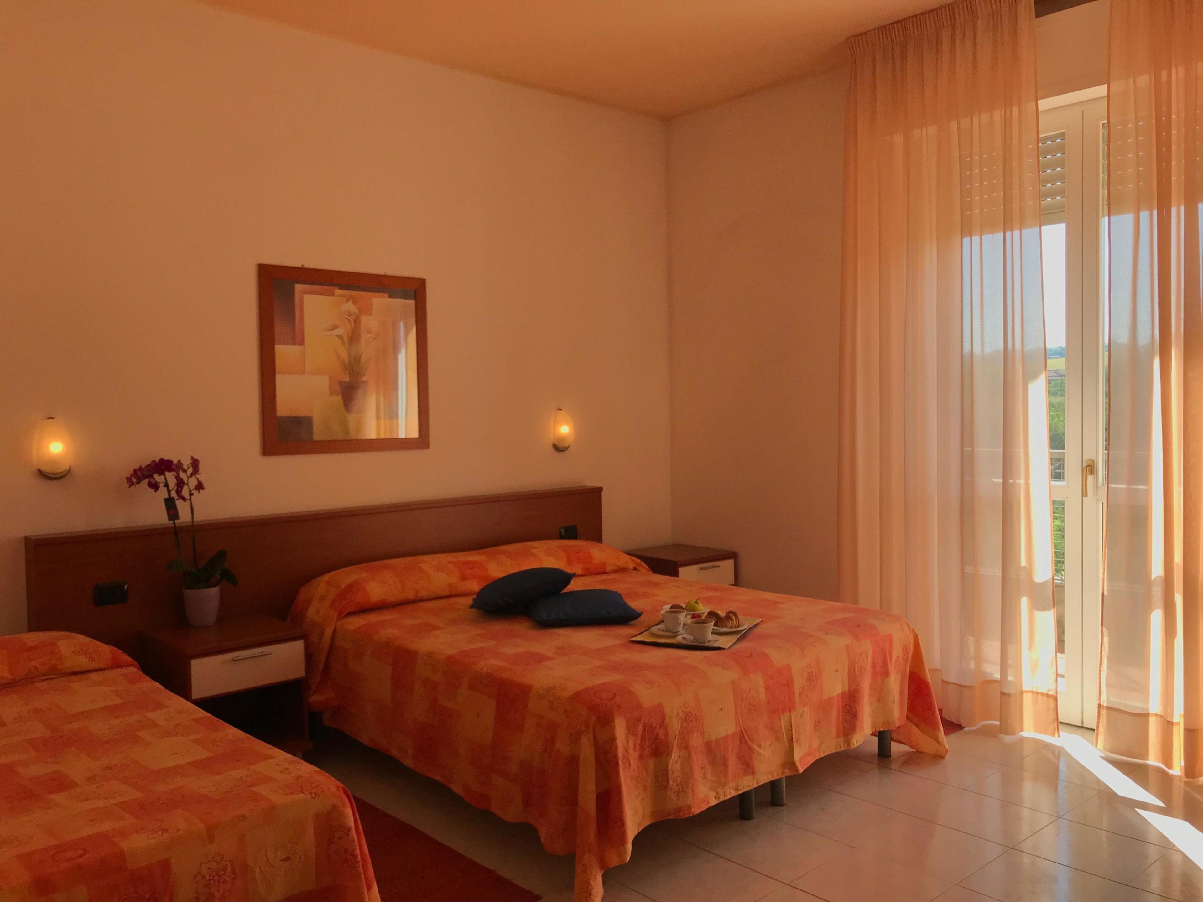 A hotel room with two beds, a nightstand, a potted plant, a painting, and a view of the outdoors through a window with sheer curtains.